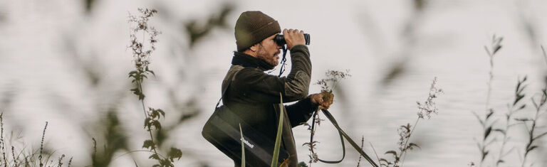 Birding With Bushnell Man standing next to a body of water looking through a pair of Bushnell Binoculars