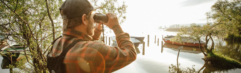 Birding With Bushnell Man next to body of water looking through Bushnell Binoculars while using a Binocular Harness